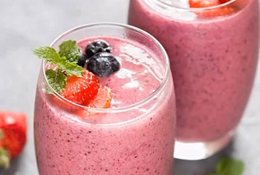 Two glasses filled with creamy pink berry smoothies topped with fresh blueberries, sliced strawberries, and a sprig of mint on a light surface.