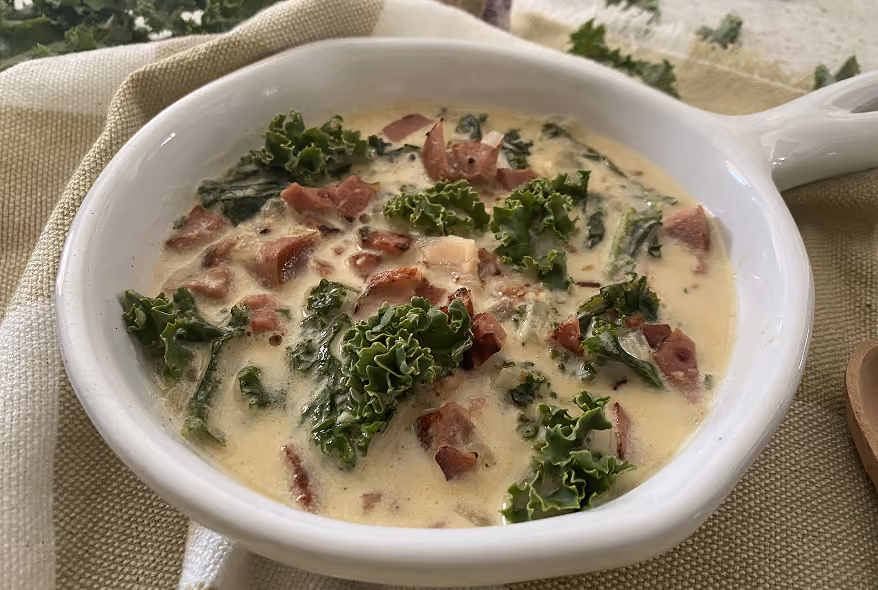 White bowl filled with creamy soup containing pieces of kale and bacon, placed on a beige and white cloth.