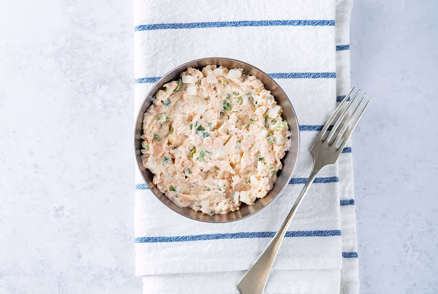 Metal bowl filled with tuna salad containing mayonnaise, chopped boiled eggs, and herbs, placed on a folded white cloth napkin with blue stripes next to a fork on a light marble surface.