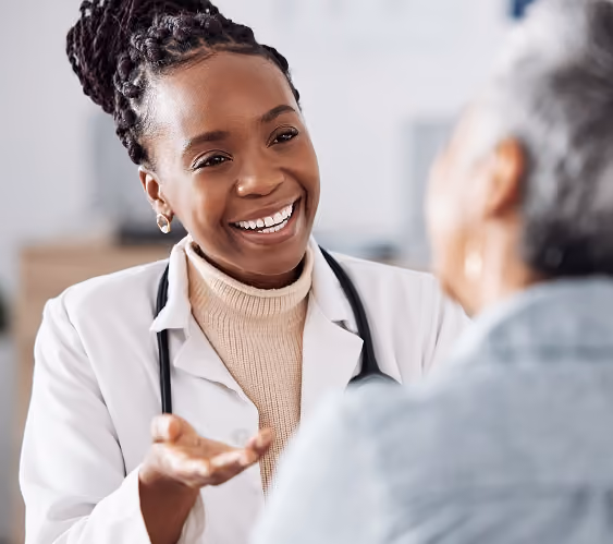 Smiling female doctor with braided hair and a stethoscope talking to an older woman in a medical office.