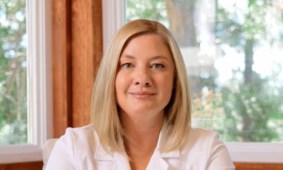 Woman with blonde shoulder-length hair, wearing a white top, sitting indoors near a window with a blurred leafy outdoor background.