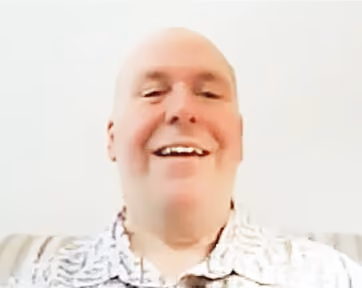 Smiling bald man wearing a patterned shirt sitting on a striped couch against a plain white background.