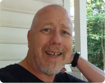 Smiling bald man with a short gray beard wearing a black shirt and a black wristwatch, posing outdoors near white siding and green trees.