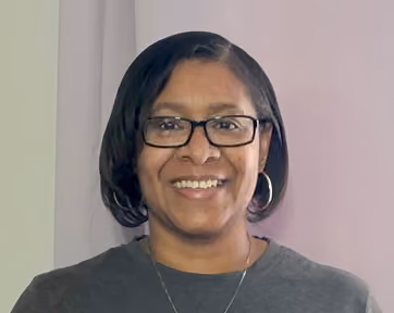 Smiling woman with short dark hair wearing glasses, silver hoop earrings, a necklace, and a dark gray shirt, standing against a light-colored background.