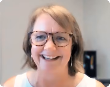 Smiling woman with light brown hair and tortoiseshell glasses wearing a white top, against a plain background with a blurred object behind her.
