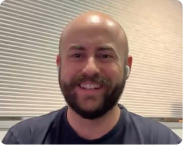 Smiling bald man with a beard wearing a dark shirt and white wireless earbuds sitting indoors in front of a window with closed blinds.