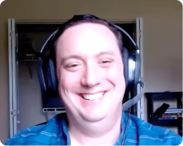 Smiling man wearing a headset and a blue striped shirt, sitting indoors with bookshelves and an open door in the background.