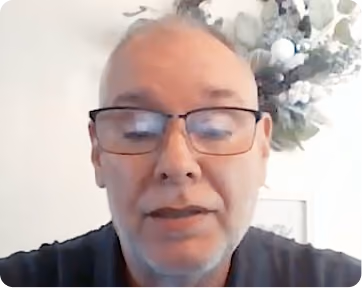 Middle-aged man with glasses and short gray hair speaking indoors with a floral decoration visible on the wall behind him.