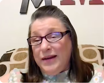 Middle-aged woman with glasses and straight shoulder-length brown hair speaks while sitting on a patterned couch with a beige pillow behind her.