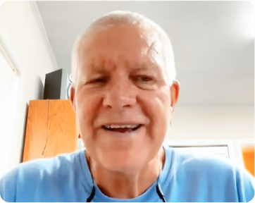 Smiling elderly man with short white hair wearing a light blue shirt and black reading glasses around his neck, sitting indoors with a wooden cabinet and window behind him.