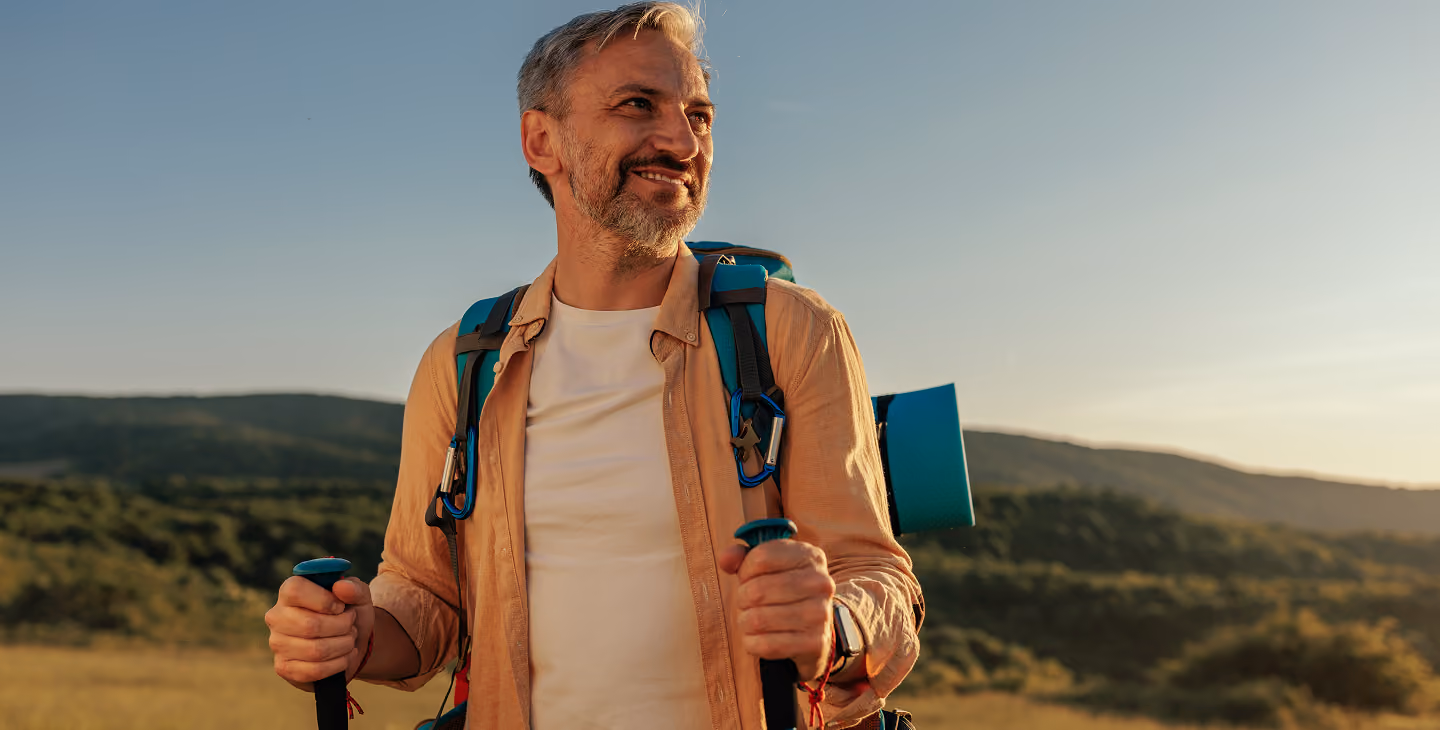 Middle-aged man with gray hair and beard smiling while hiking outdoors, wearing a backpack and holding trekking poles with hills and clear sky in the background.