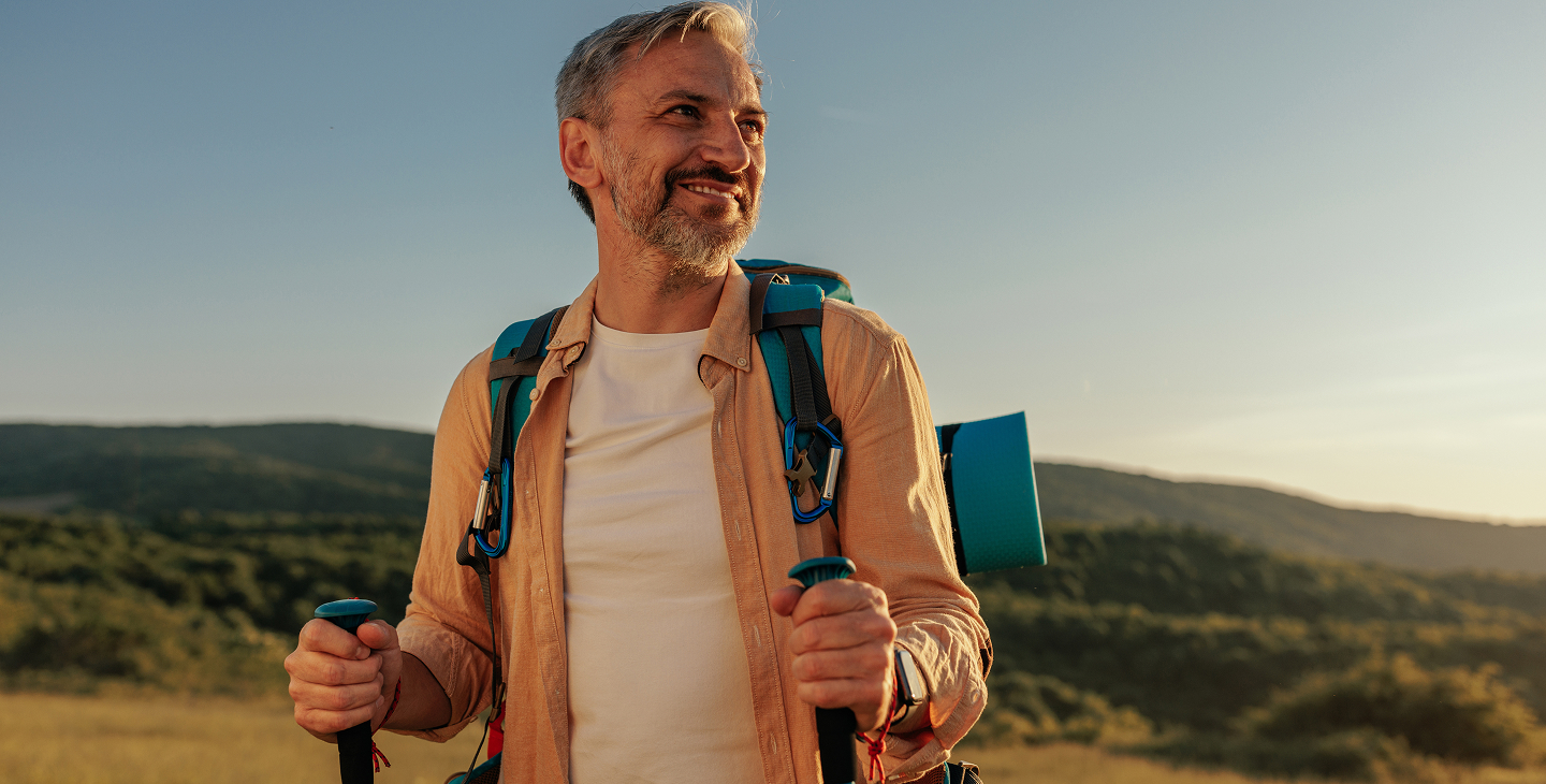 Middle-aged man with gray hair and beard smiling while hiking outdoors, wearing a backpack and holding trekking poles with hills and clear sky in the background.