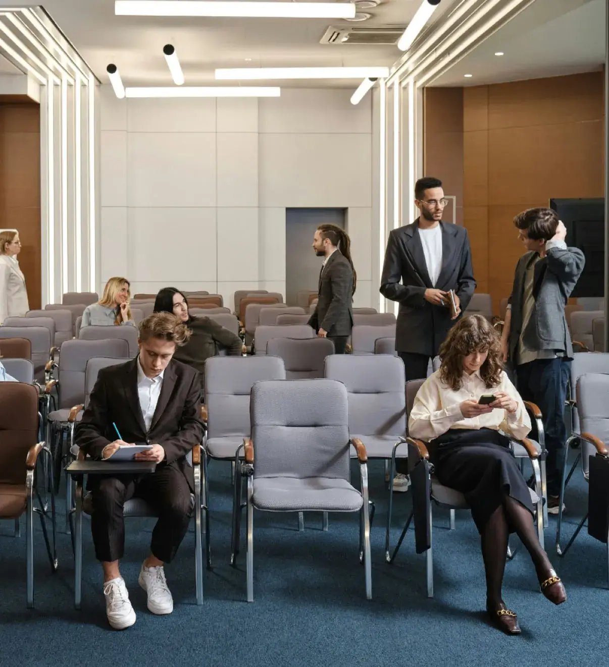 People gathering and preparing for a session in a modern conference room with rows of chairs and bright overhead lighting.