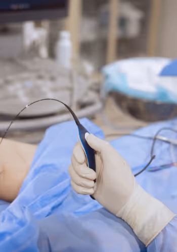 Medical professional wearing gloves holds a thin catheter-like device near a patient's covered arm in a clinical setting.