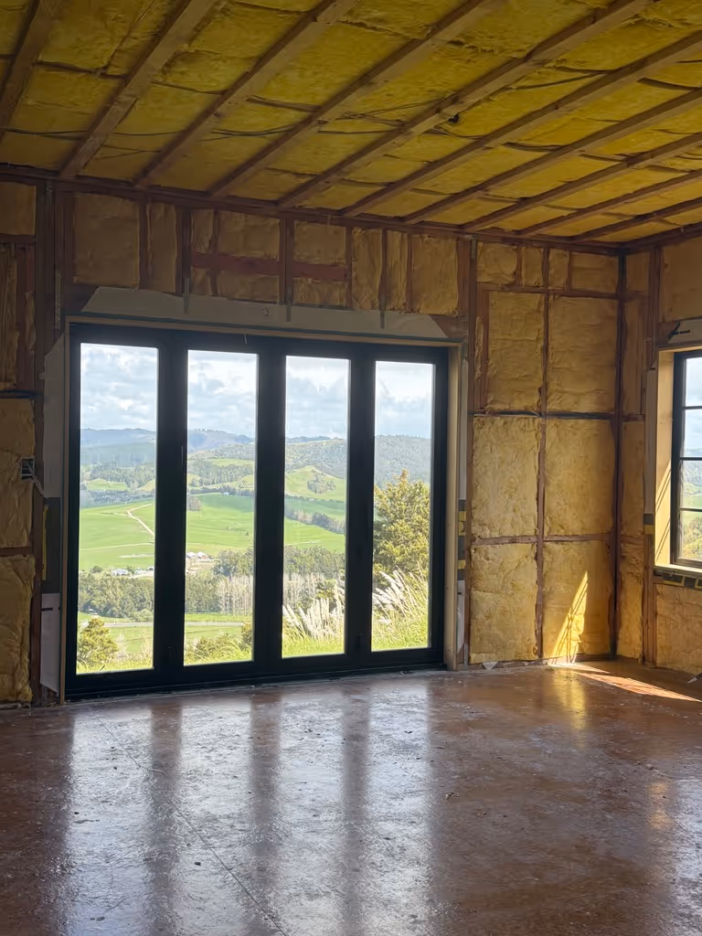 Interior of a room under construction with exposed insulation on walls and ceiling and large glass doors showing a green landscape outside.