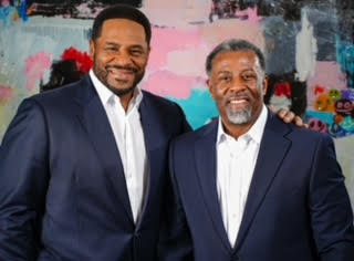 Two men smiling in dark suits and white shirts standing in front of a colorful abstract wall.