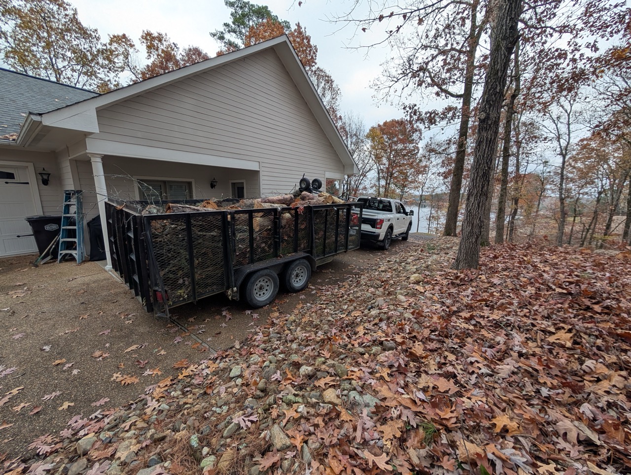 White truck towing a black trailer loaded with cut logs parked next to a house in autumn.
