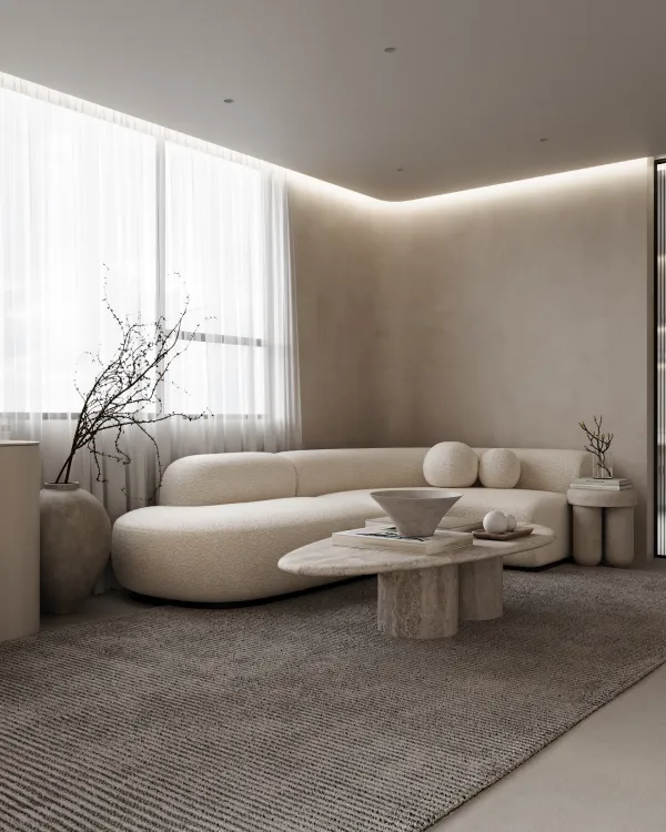A serene lounge area with a large curved sofa on a textured rug, centered around a rustic stone coffee table. The room features soft natural lighting from a window covered with sheer curtains and is decorated with minimalist vases and an abstract branch arrangement.
