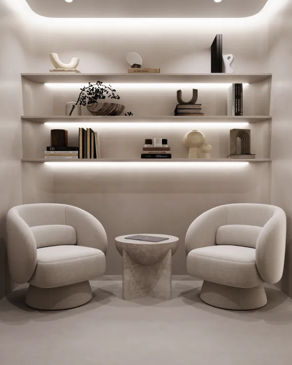 A cozy waiting area in a spa with two plush, curved chairs facing each other over a small stone table. Above, shelves display various decorative items and books under soft, ambient lighting.