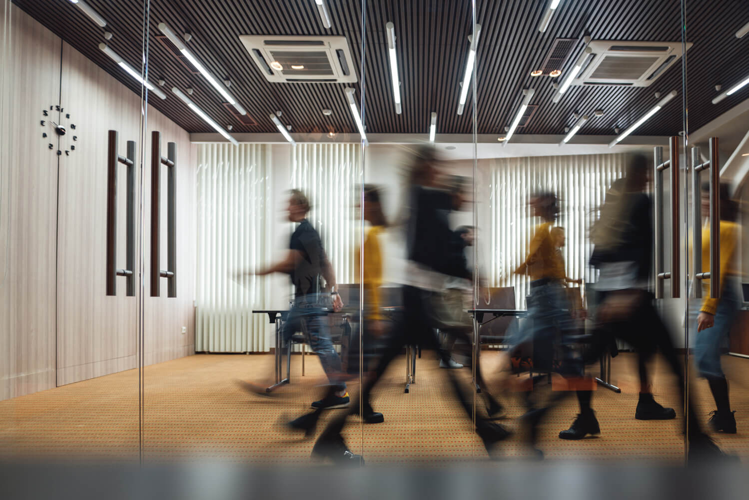 Blurred motion of people walking through a modern office space with glass walls and a wooden ceiling, emphasizing movement and activity.