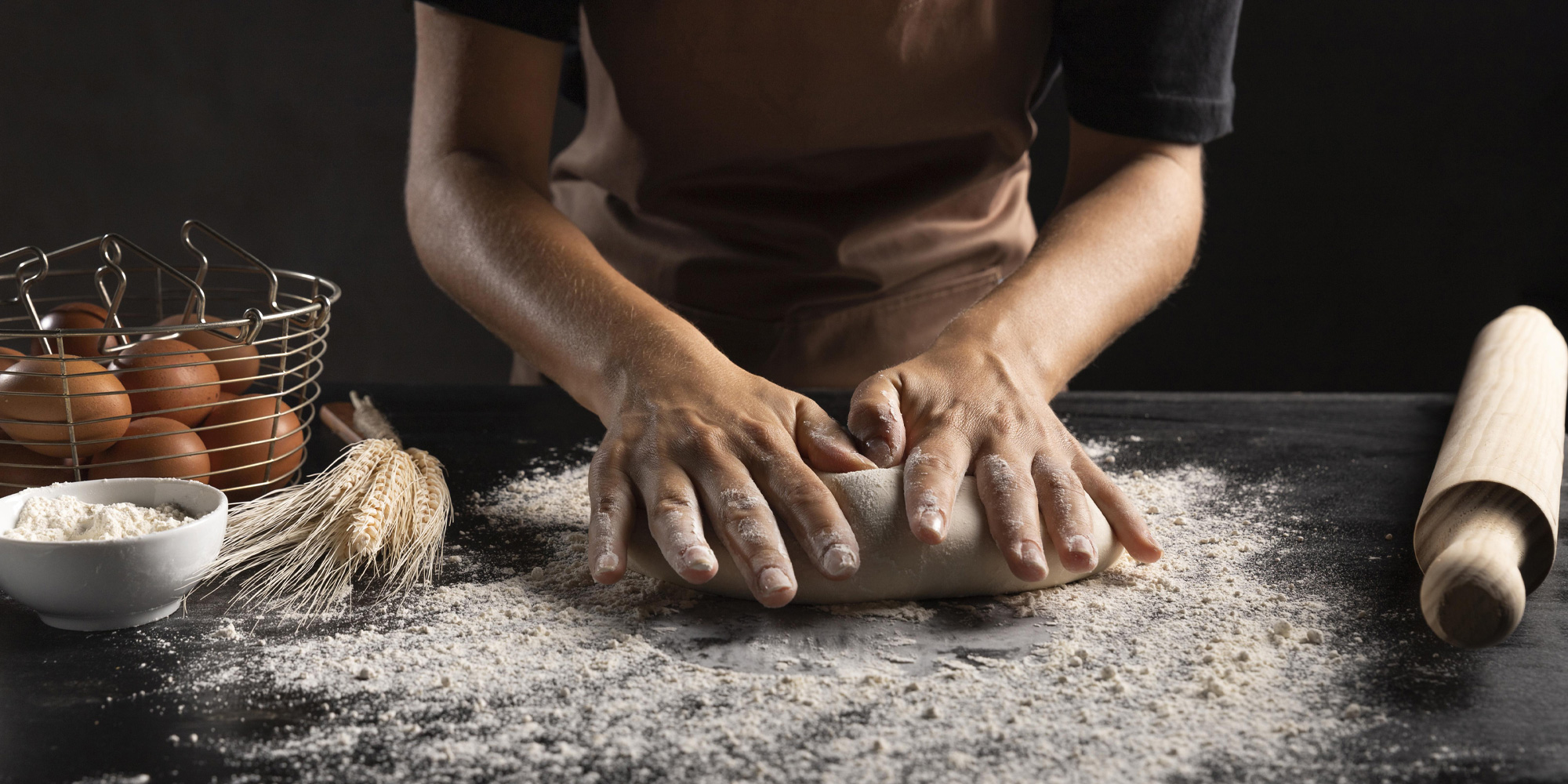 person kneading dough on a black table dusted with flour. wheat, eggs, flour, and wooden roller in the background