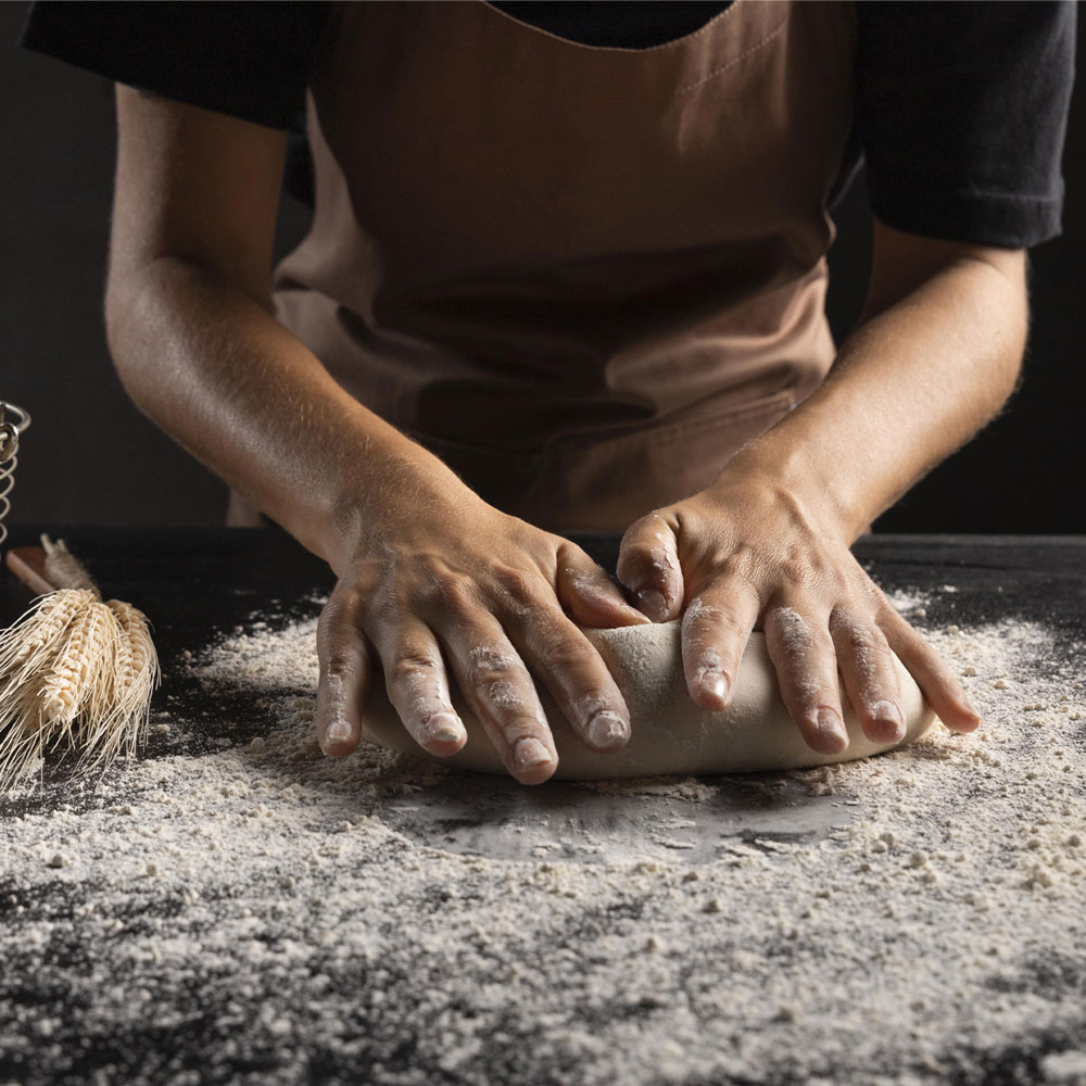 person kneading dough on a black table dusted with flour. wheat, eggs, flour, and wooden roller in the background