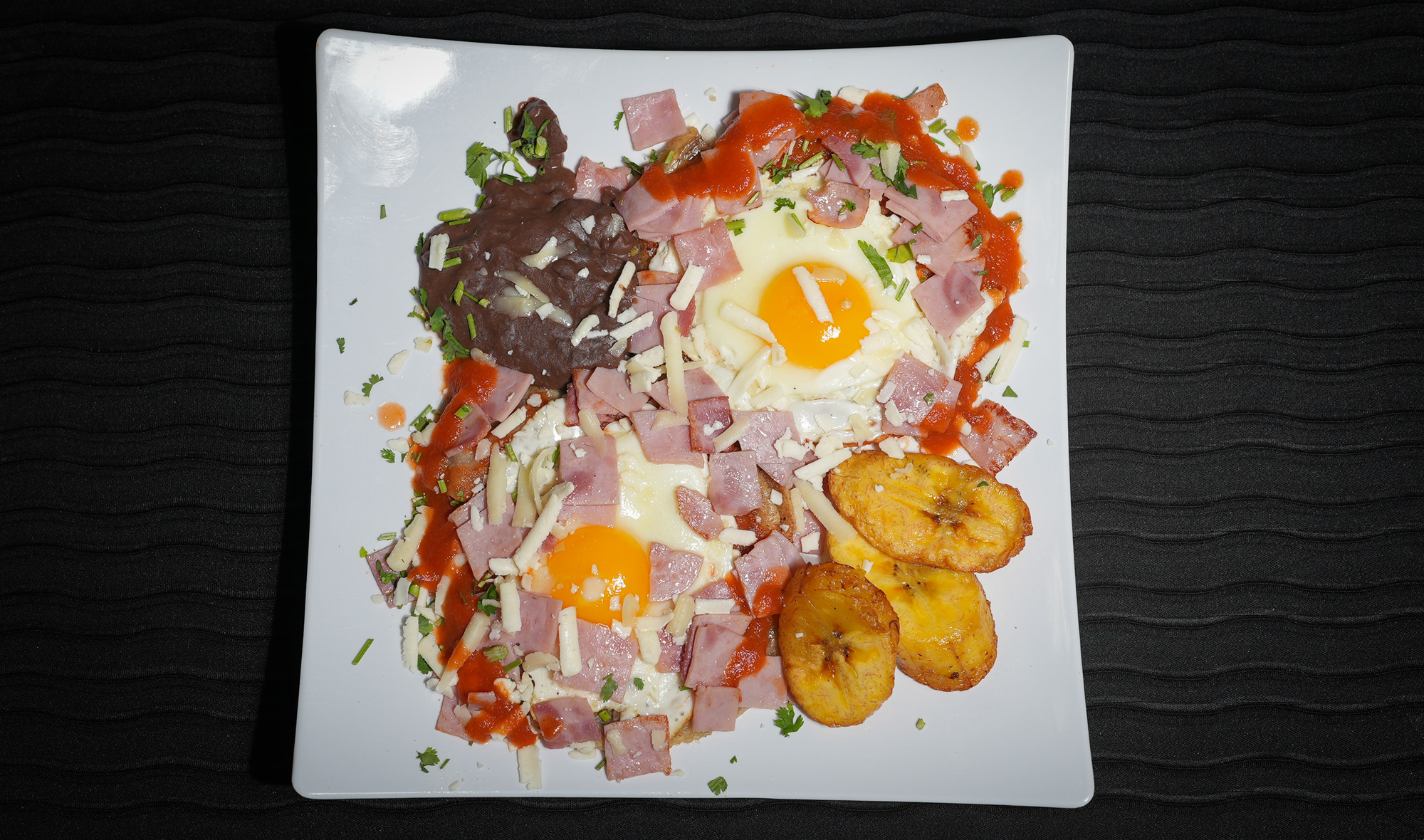 two sunnyside eggs with red salsa, scattered ham and refried beans with plantains