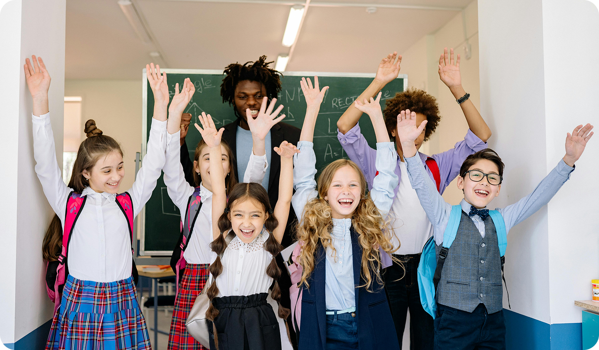 Diverse group of smiling schoolchildren with backpacks raising their hands excitedly in a classroom hallway in front of a chalkboard.