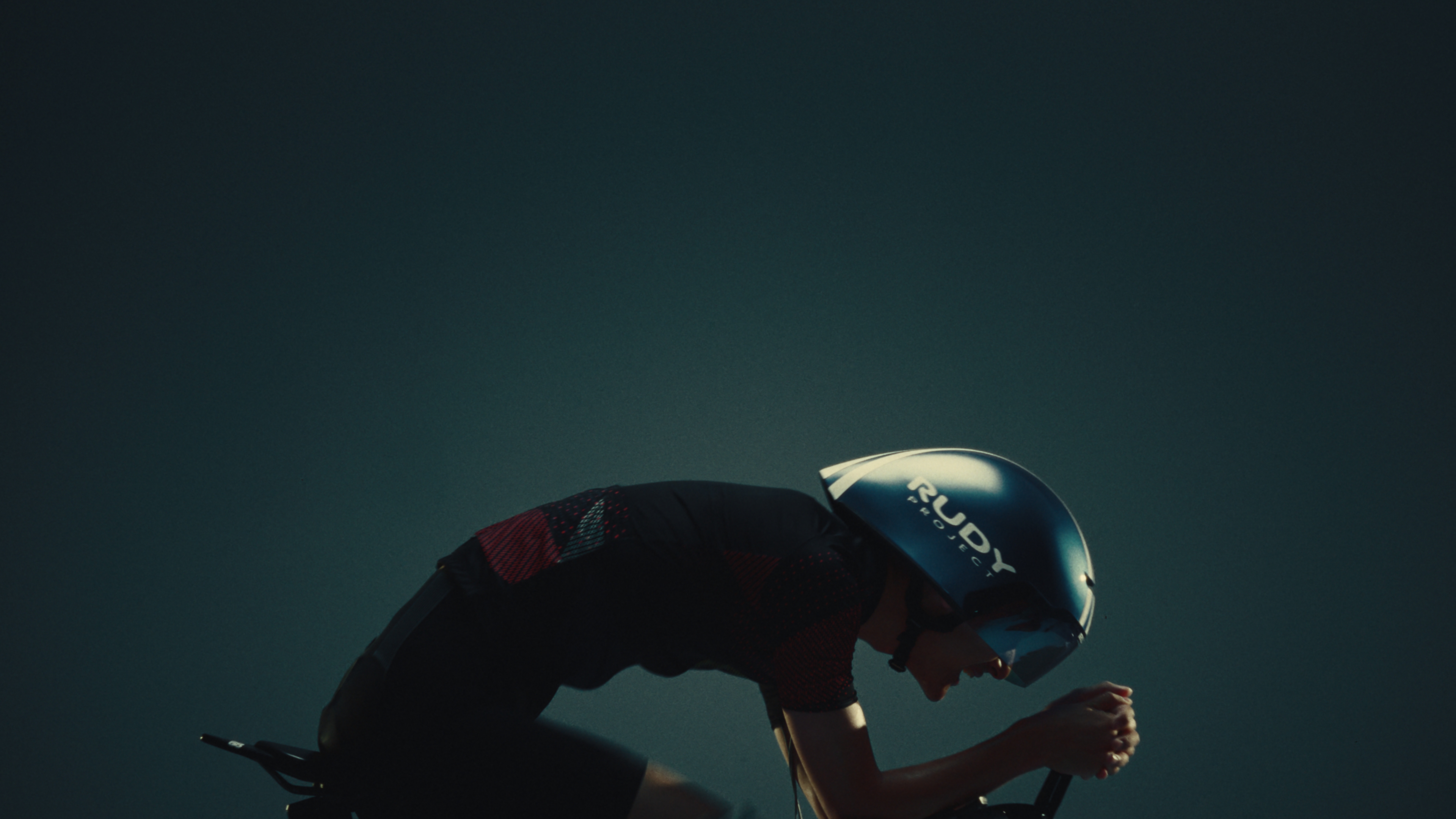 Cyclist in a Rudy Project time trial helmet tucked low in an aero position against a dark teal backdrop