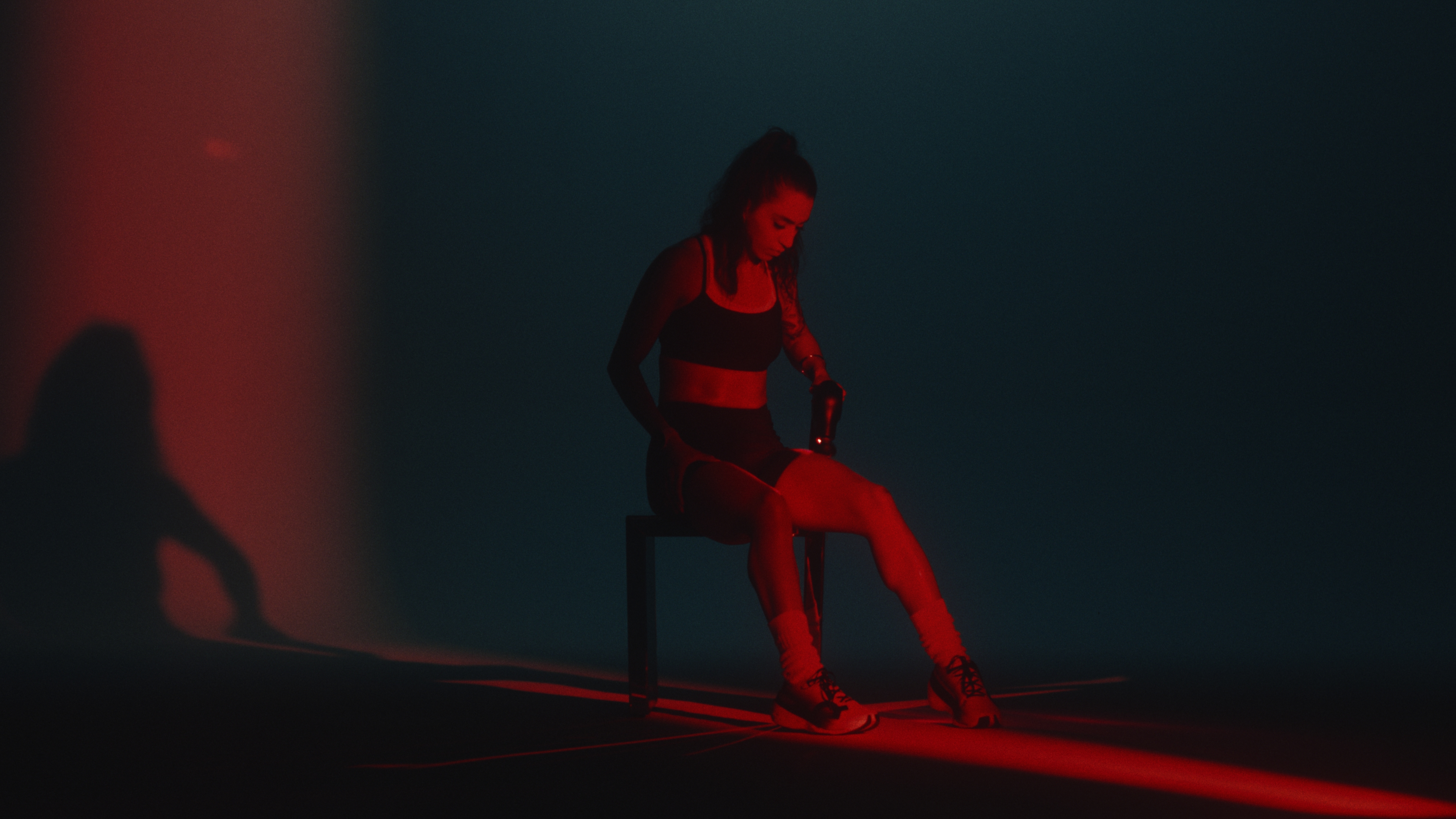 Athlete seated in red light using a Theragun on her leg, her shadow cast on the wall behind her