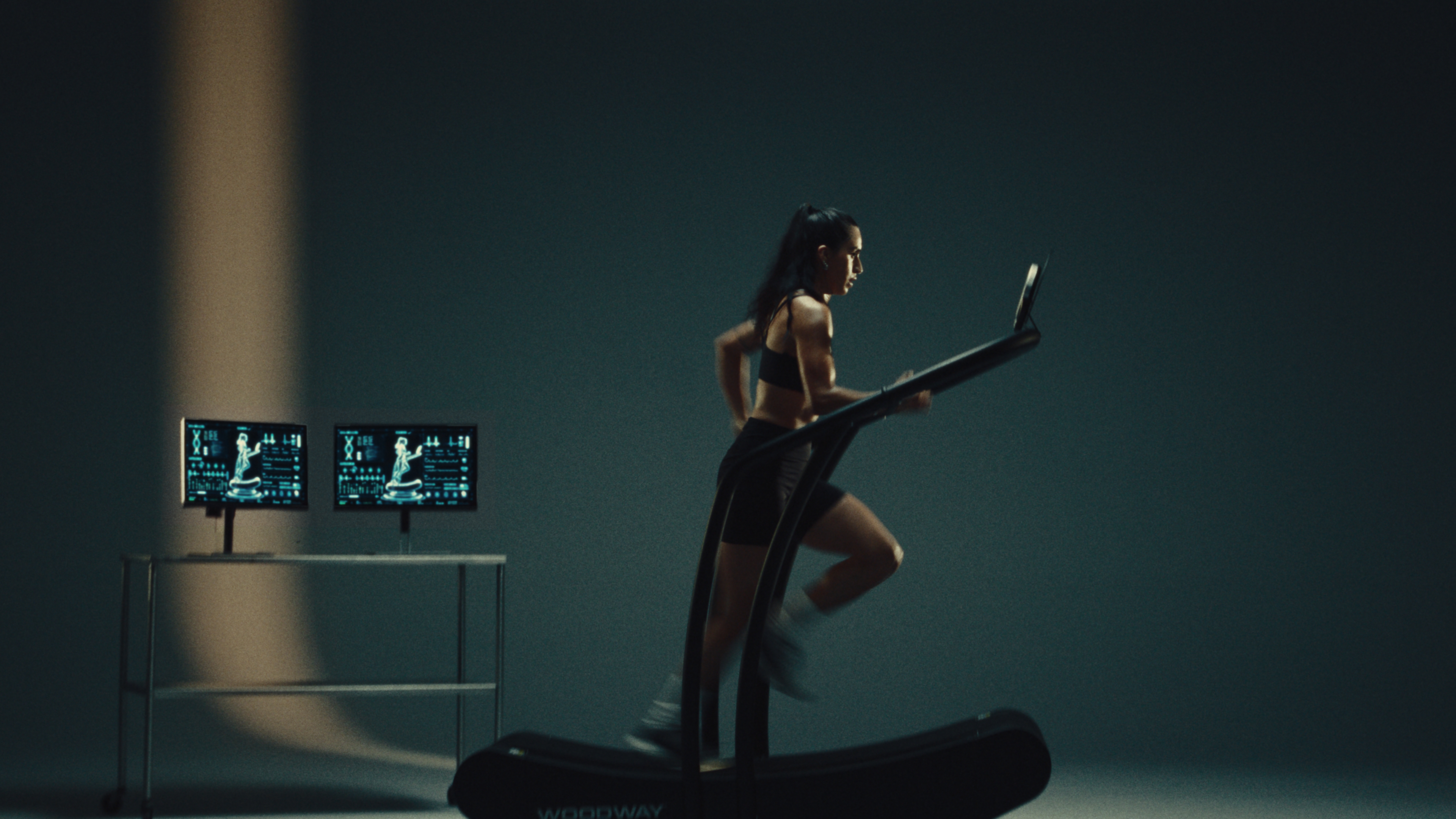 Runner on a Woodway curved treadmill beside two monitors displaying biometric data on a dark stage