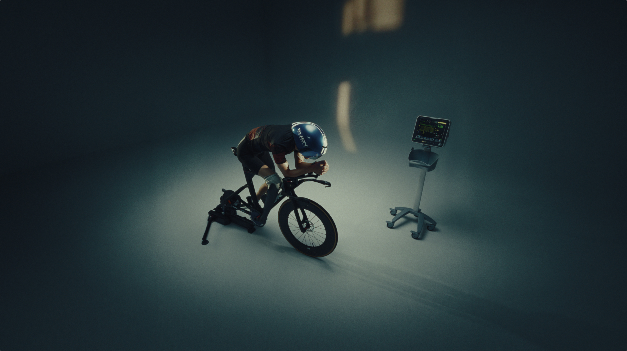Wide view of a cyclist on a stationary trainer beside a biometric monitor on a dark stage