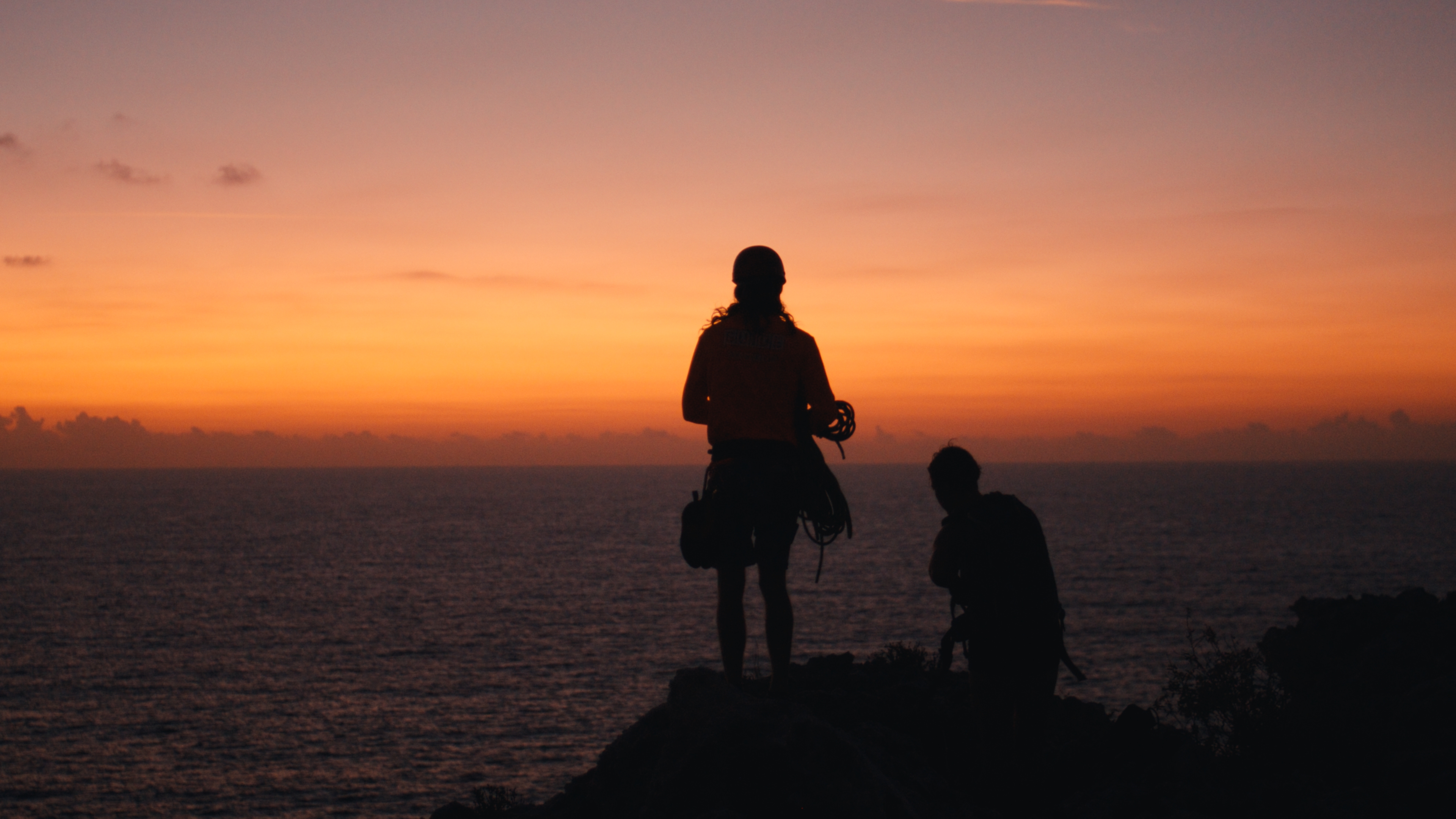 Two filmmakers silhouetted on a sea cliff at sunset on Cayman Brac
