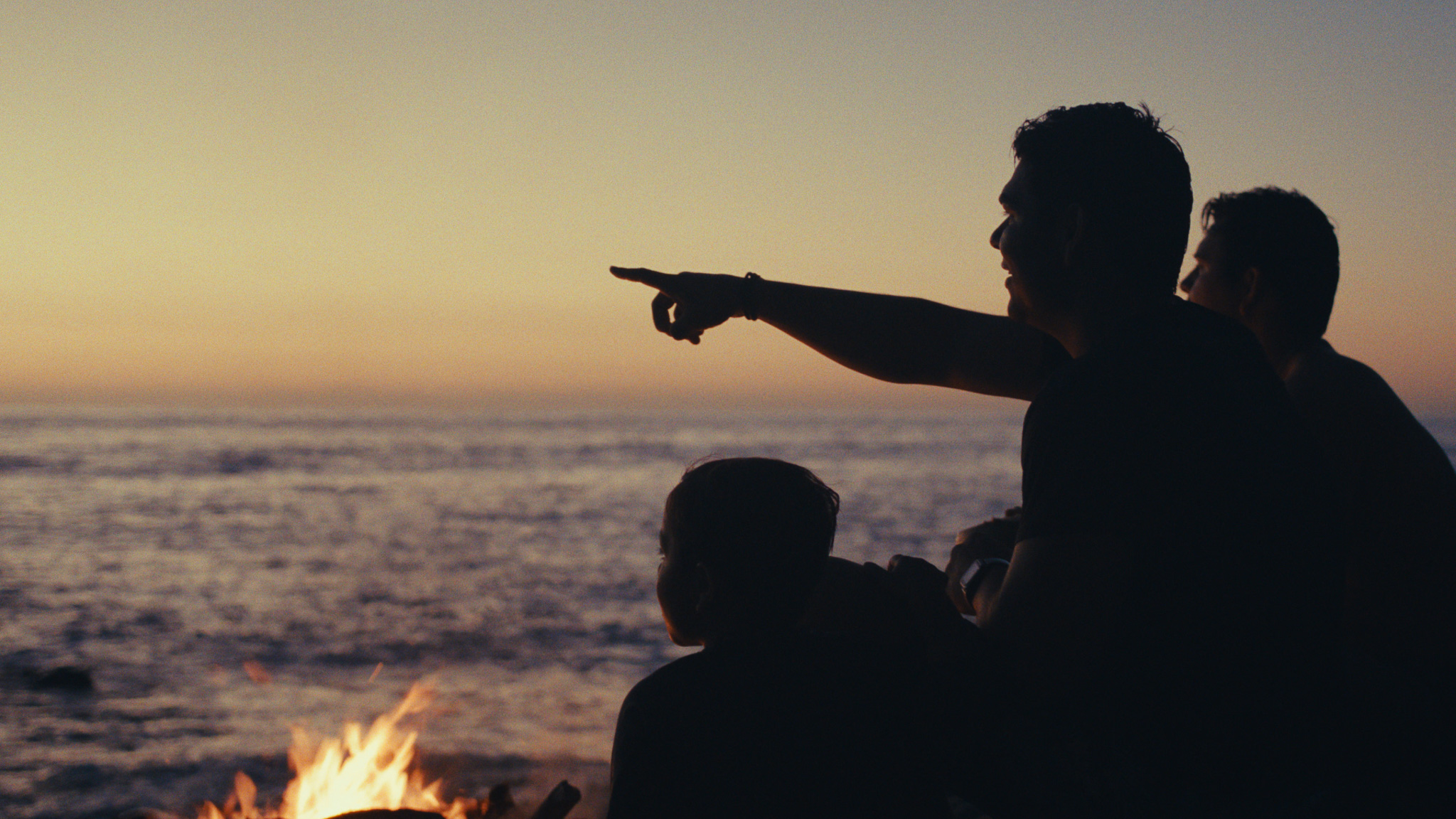 Men pointing out to sea at dusk beside a fire