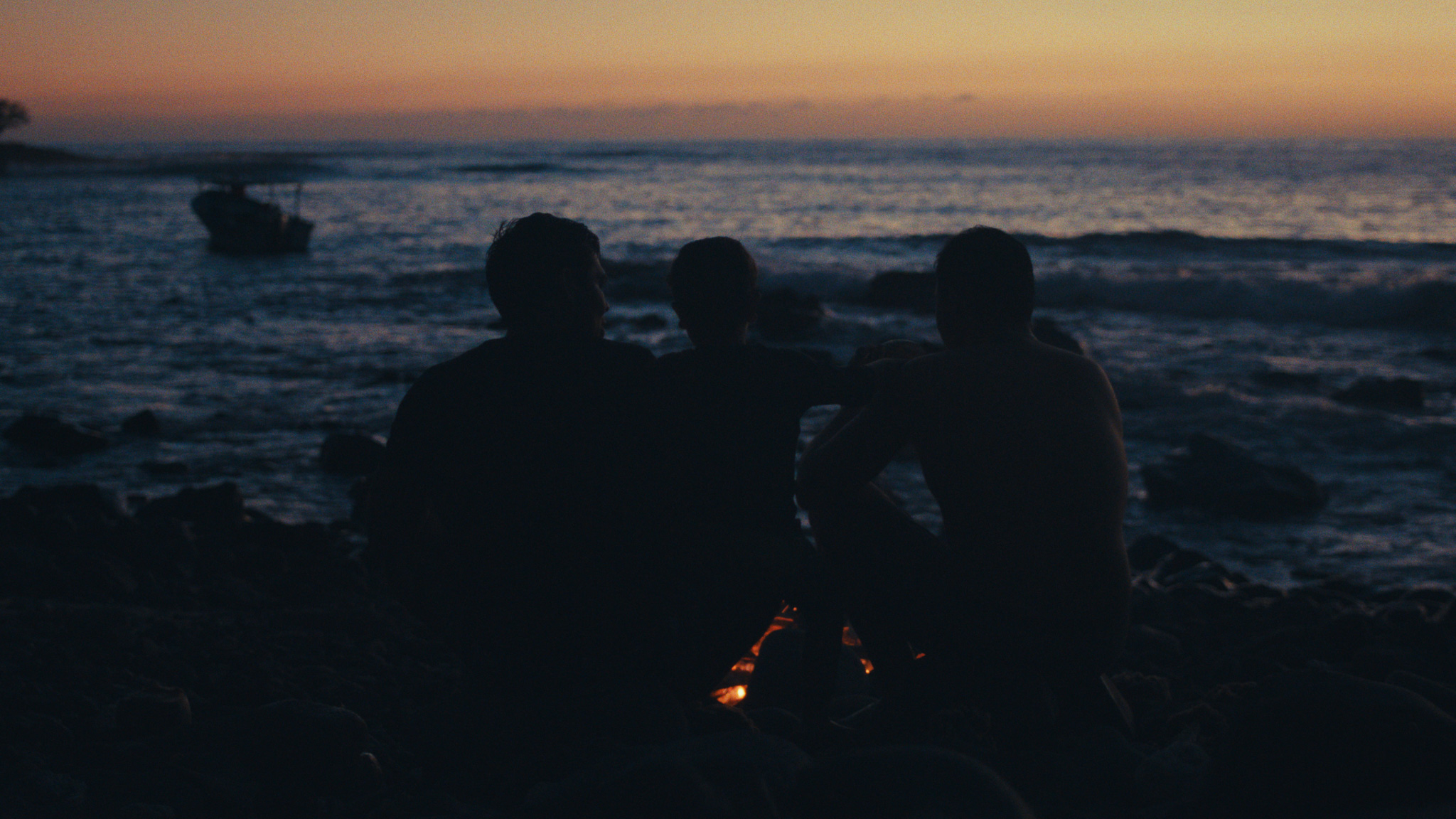 Three men watching the sea at sunset with a boat offshore