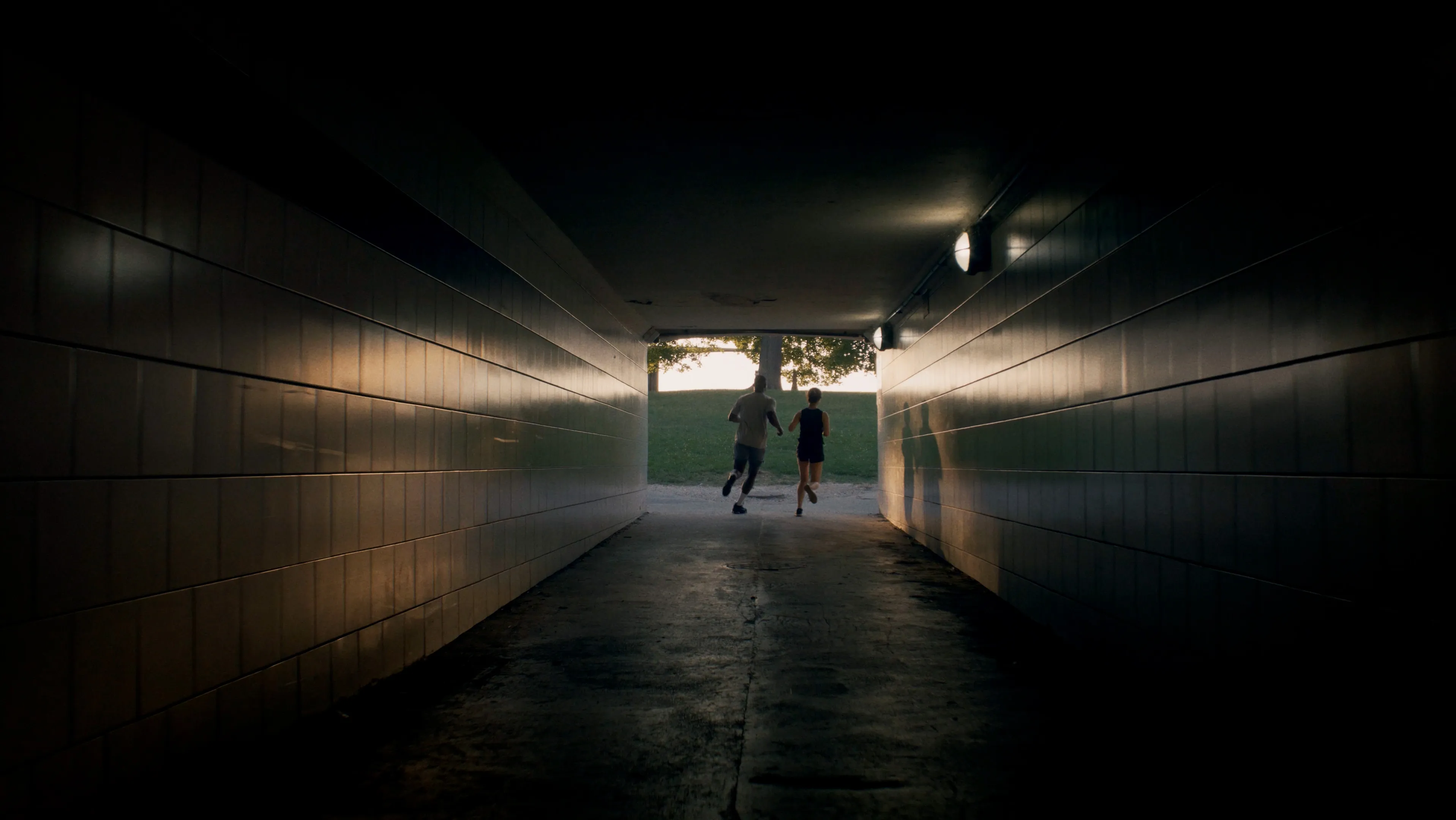 Two runners moving through a dark pedestrian tunnel toward the light