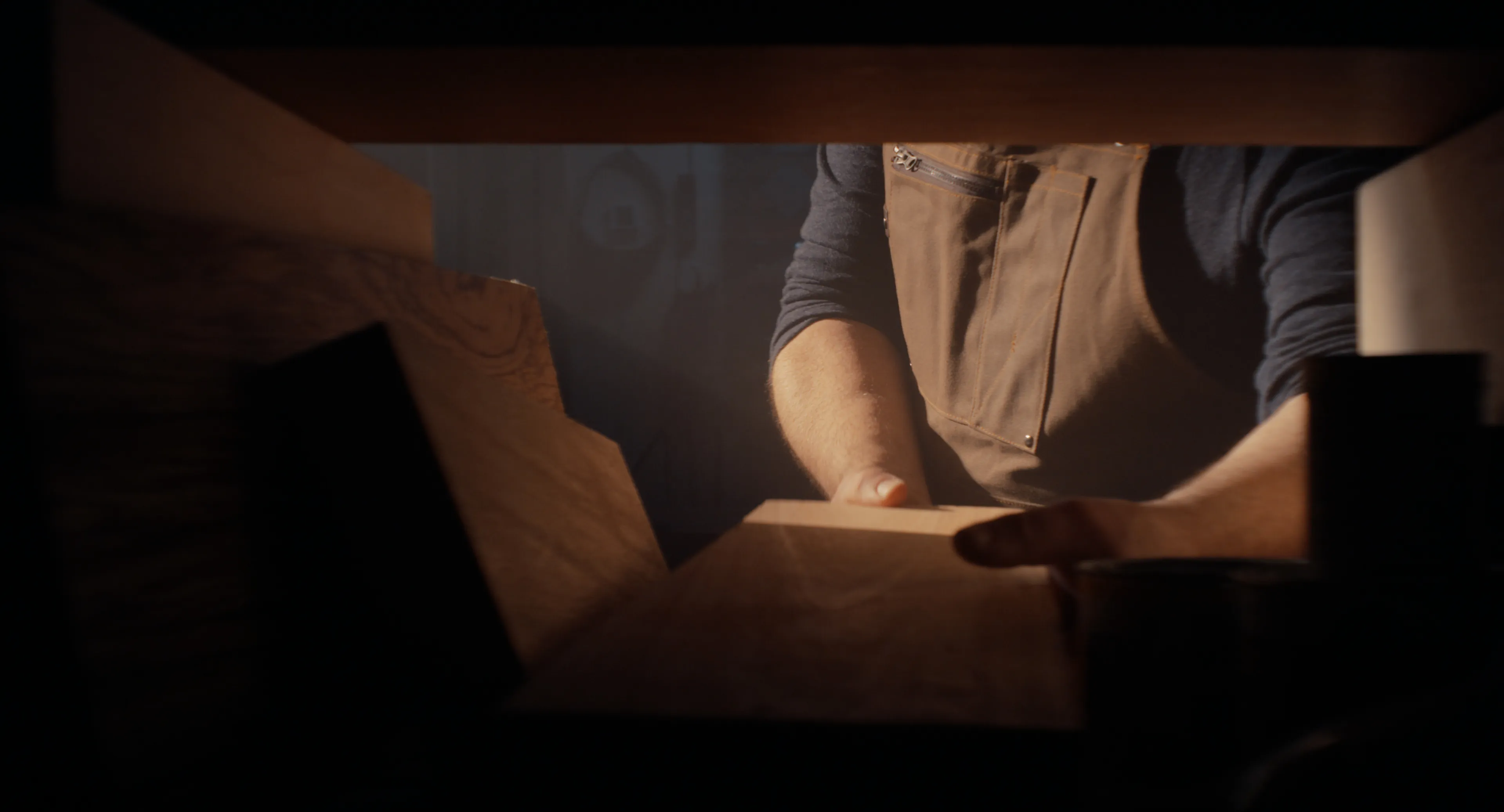 Luthier leaning over a slab of tonewood between stacked lumber in warm light
