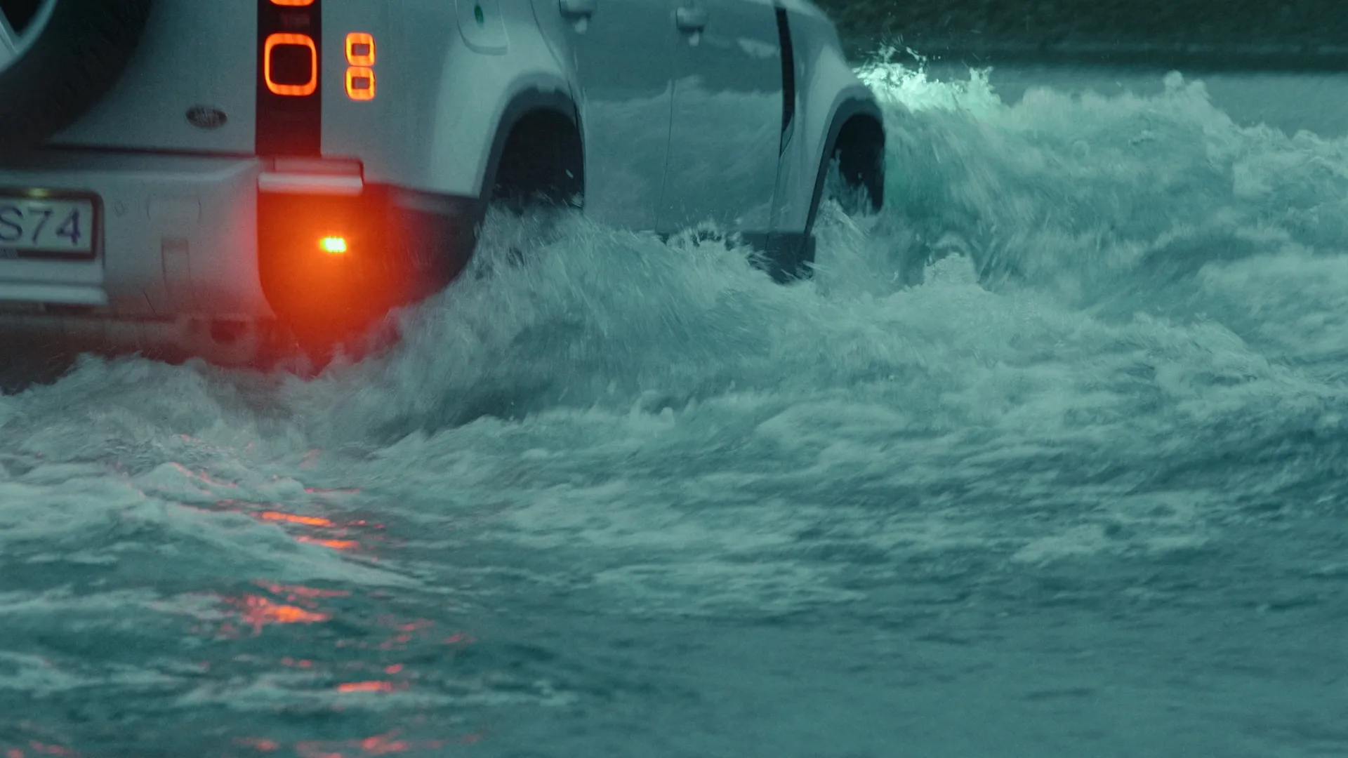 Rear of the Defender crossing a churning river with tail lights glowing