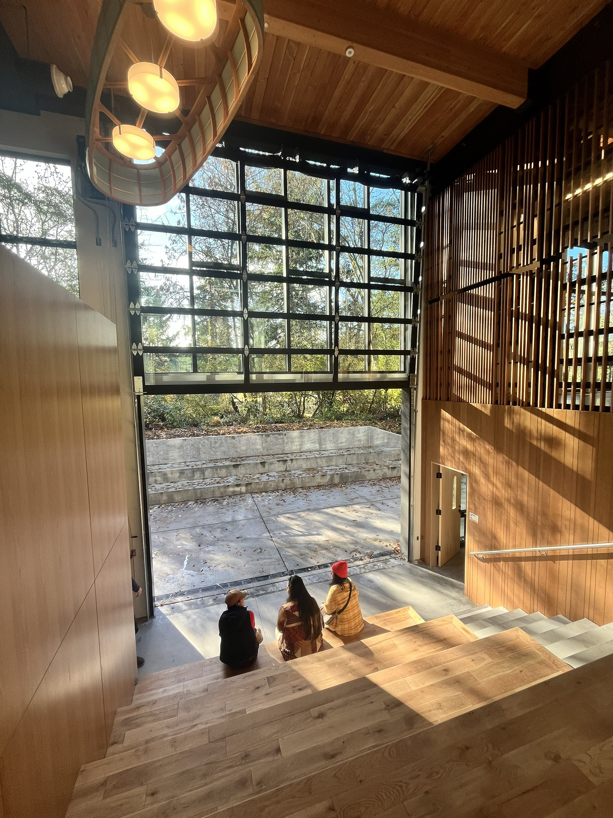 Wood stadium stair with 3 people sitting at the bottom of the stairs looking out an open garage door toward trees