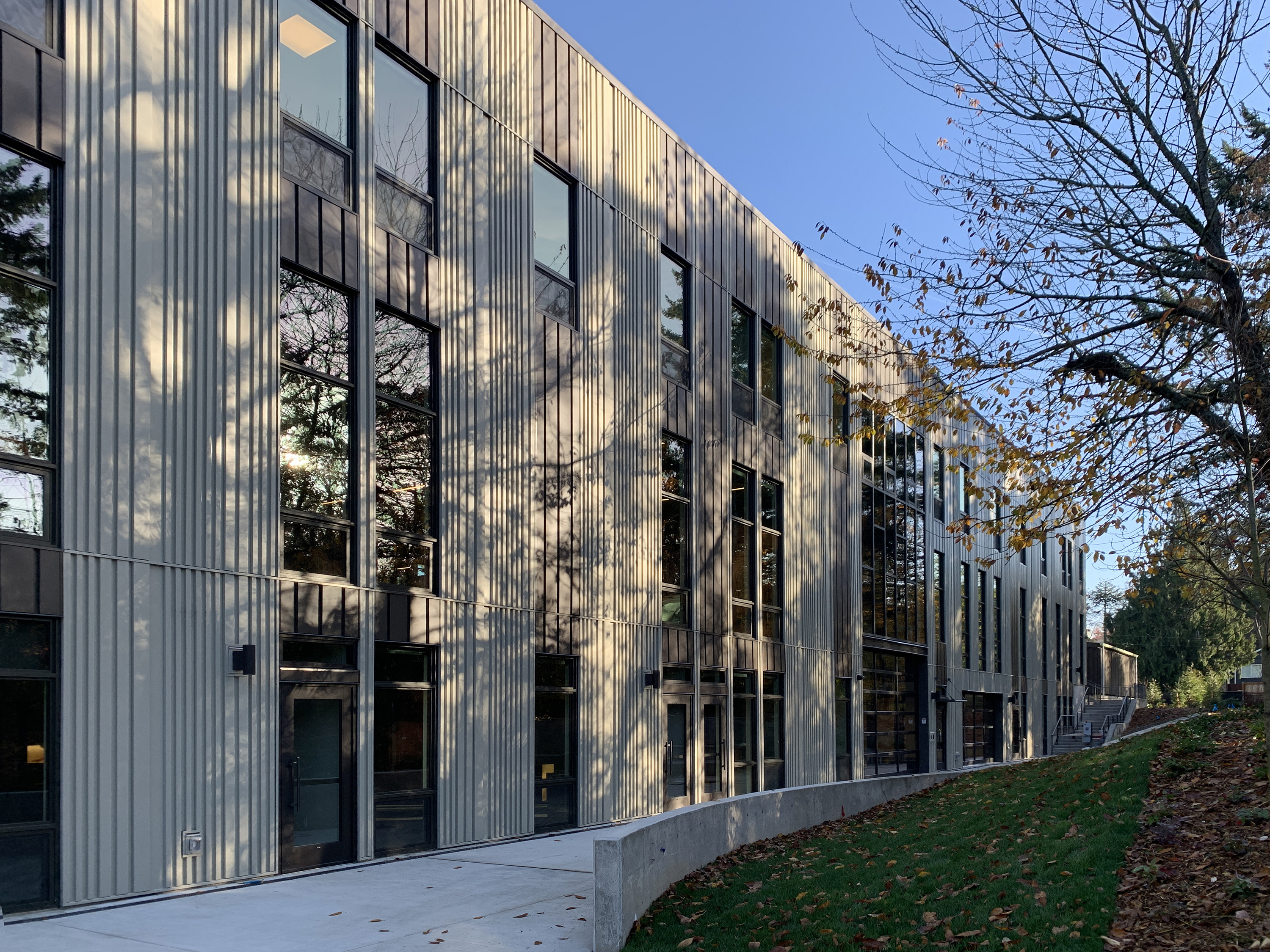 rectangular building with vertical offwhite metal siding. Blue sky and sun and tree shadows on the building