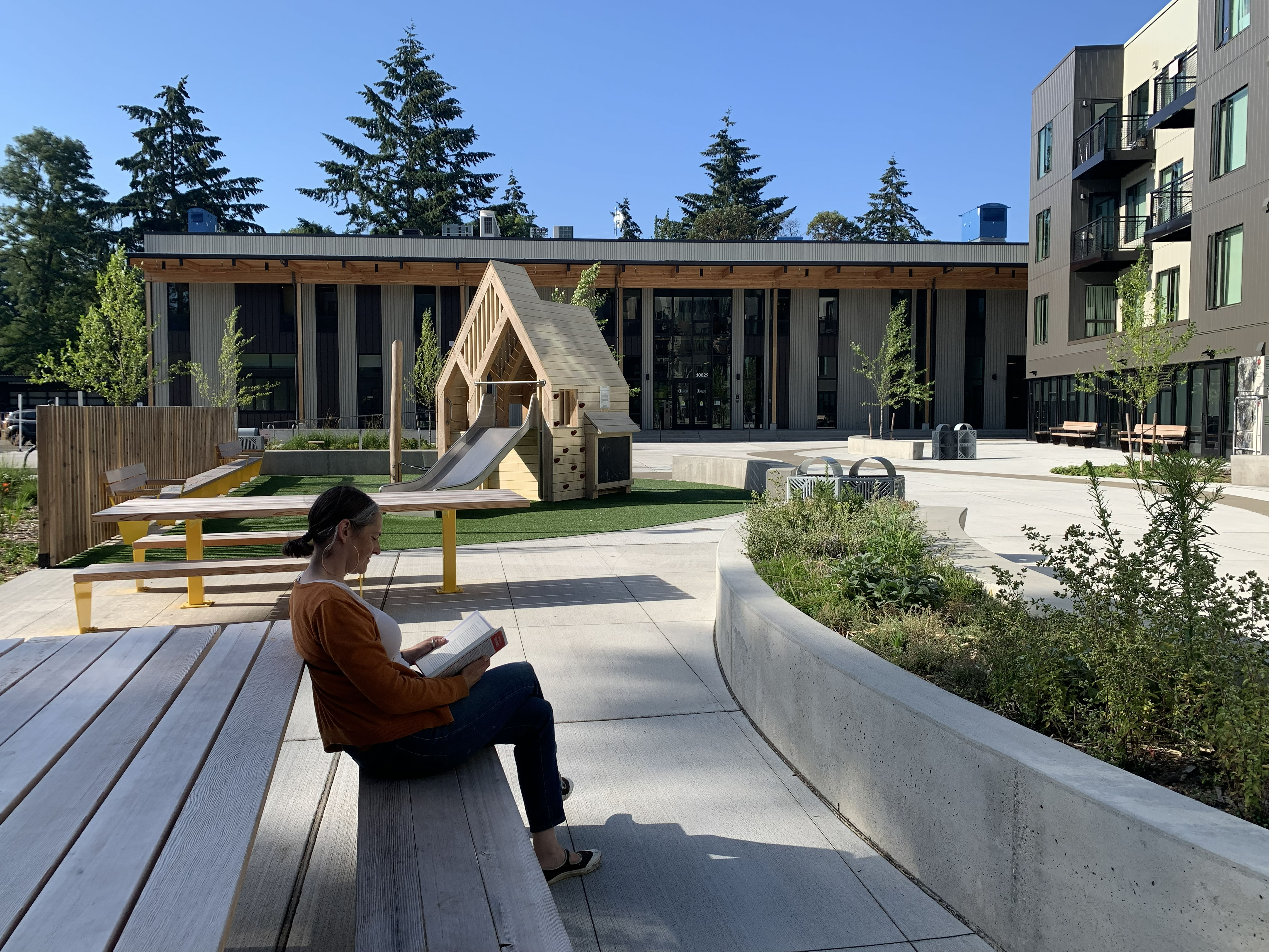 rectangular community building in the background, house shaped playground in the middle ground, woman reading at picnic table in the foreground