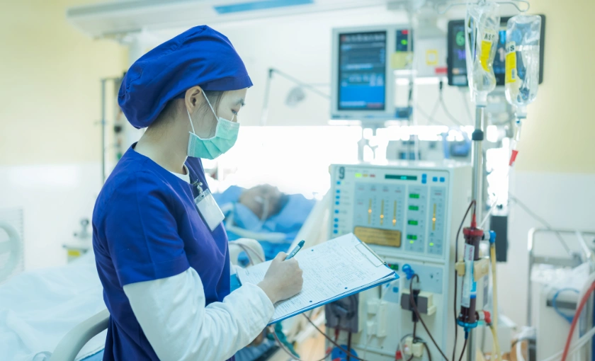 Nurse wearing personal protective equipment monitoring a dialysis machine and reviewing treatment settings during a dialysis session.