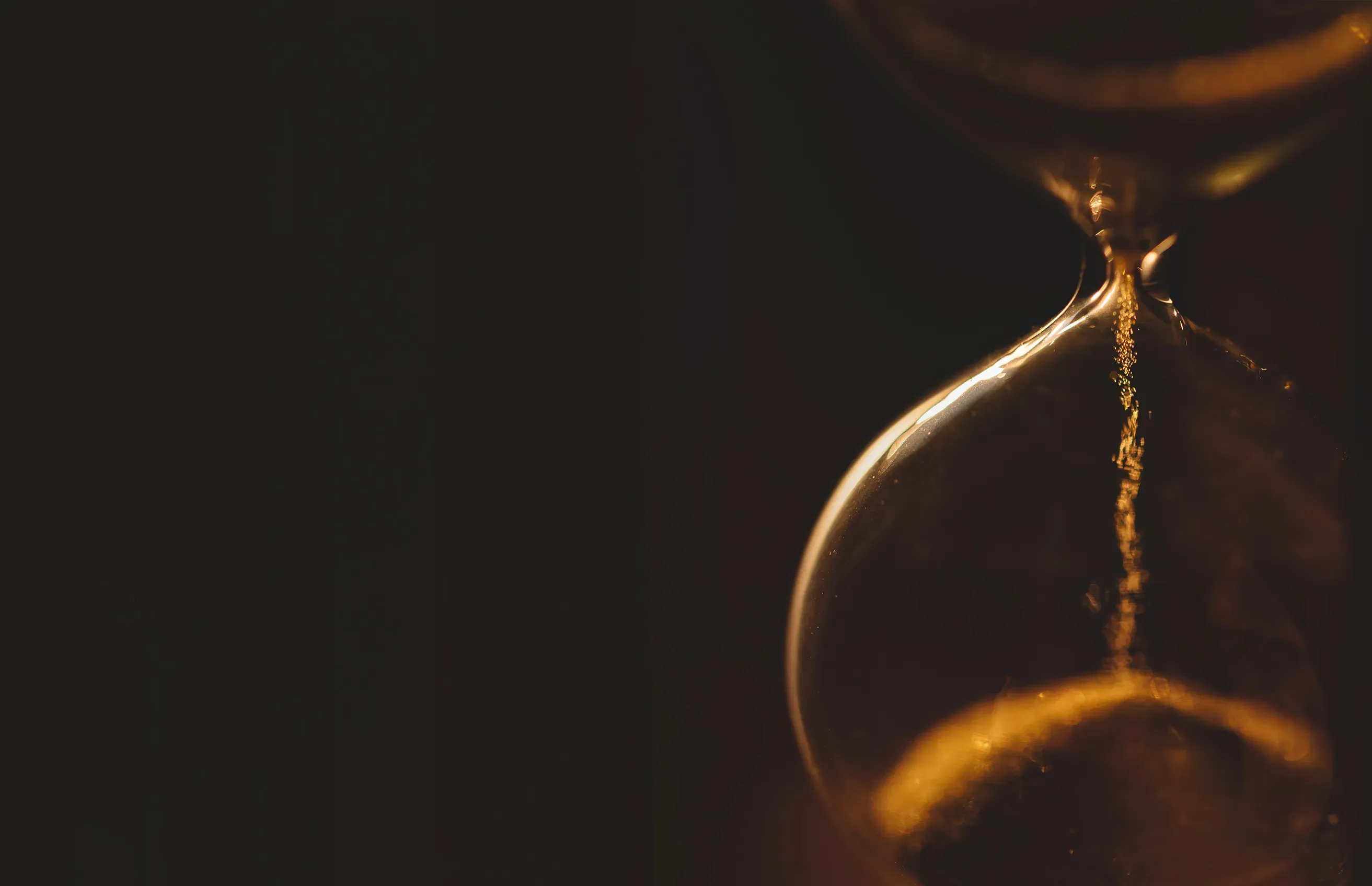 Close-up of an hourglass with sand flowing through the narrow middle section against a dark background.