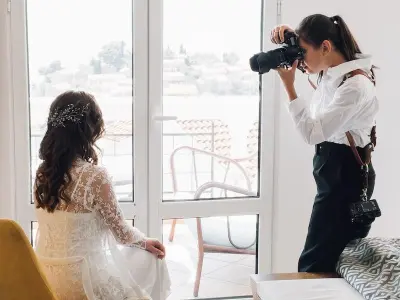 Wedding videographer filming a bride in a lace wedding dress near a window during bridal preparations, capturing natural light and candid pre-ceremony moments.