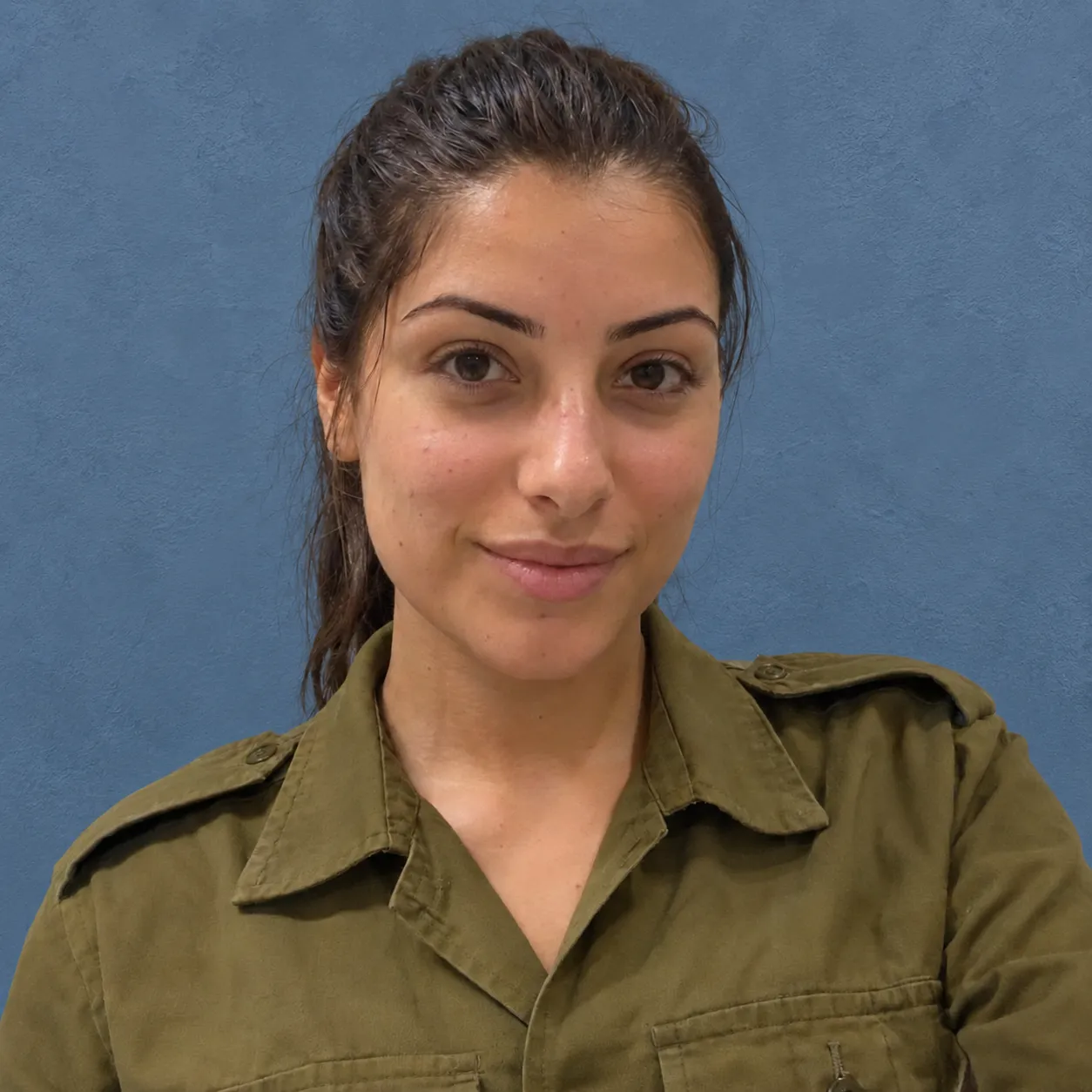 Young woman with dark hair tied back, wearing an olive green buttoned shirt, smiling slightly against a plain blue background.