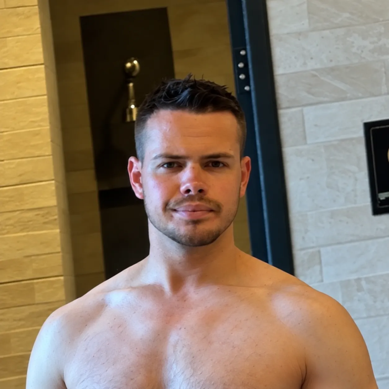 Shirtless young man with short dark hair and light facial hair standing indoors against a tiled wall with a dark framed mirror behind him.