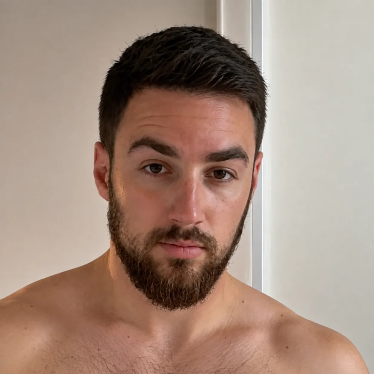 Portrait of a shirtless man with short dark hair and a full beard, looking directly at the camera with a neutral expression against a plain light background.
