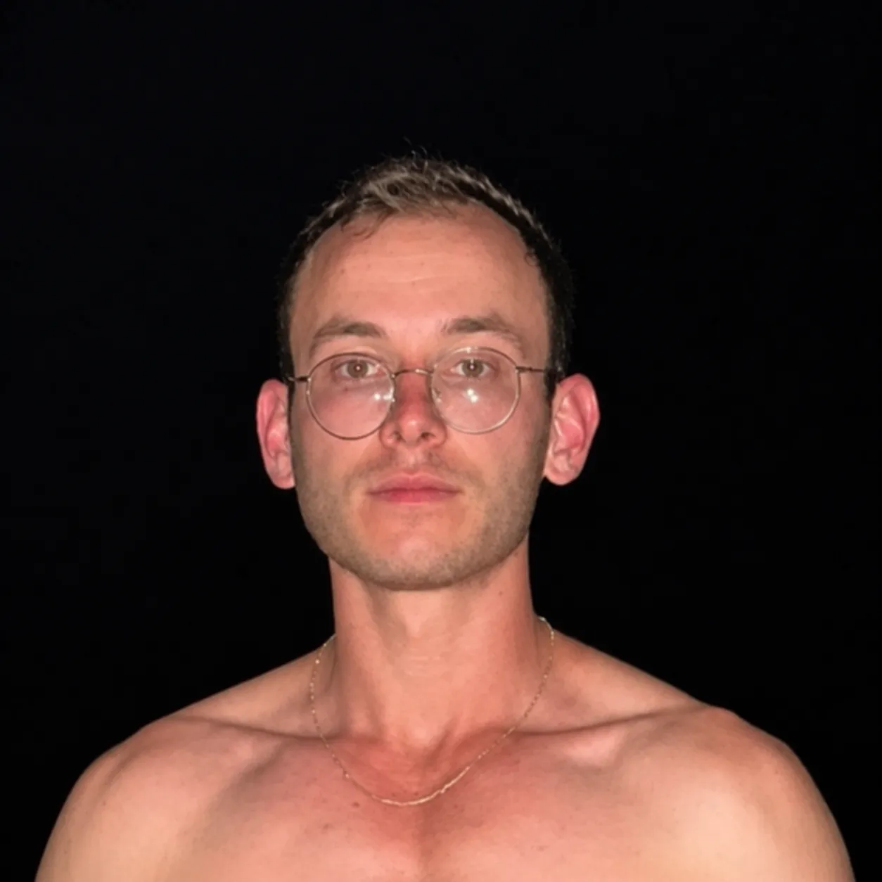 Shirtless man with short light brown hair, wearing round eyeglasses and a thin chain necklace, against a black background.
