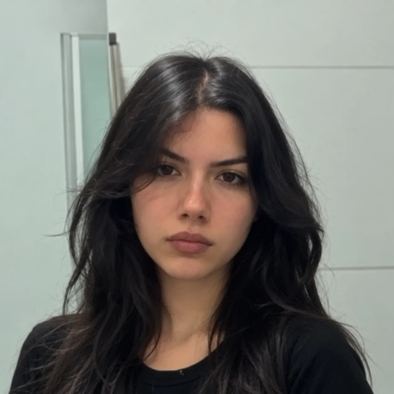 Portrait of a young woman with long dark wavy hair and a neutral expression wearing a black shirt against a plain light background.