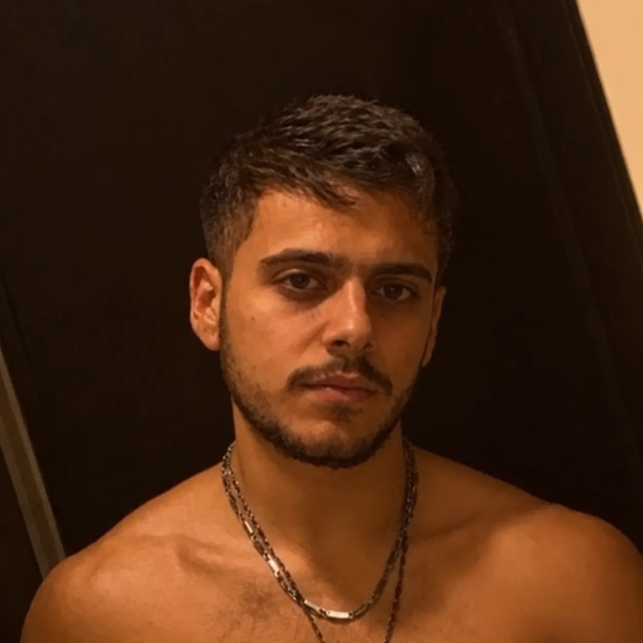 Young man with short dark hair and a trimmed beard, wearing two silver chains, posing shirtless against a dark background.